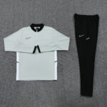 Nike athletic tracksuit top and matching pants laid flat on a gray carpet; light gray pullover with black collar and zip, black joggers with white swoosh detail.