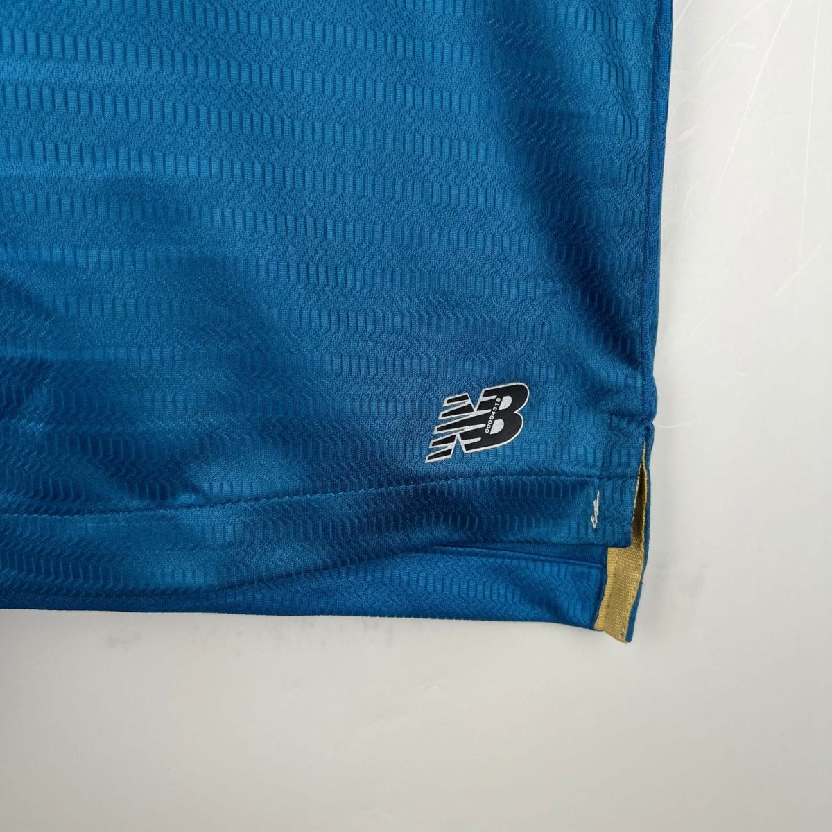 Close-up of blue fabric with a subtle striped pattern and a white New Balance logo in the bottom-right corner, yellow strap visible on the edge.