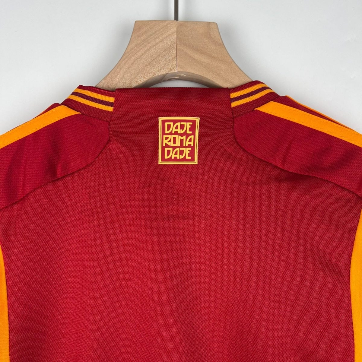 Red sports jersey on a wooden hanger, with yellow stripes along the shoulders and a yellow logo patch reading DAJE ROMA DAJE on the back neck area.