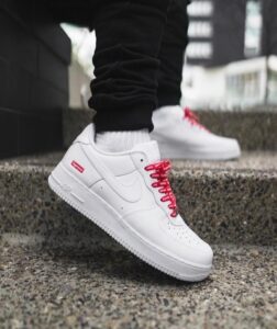 White Nike Air Force 1 sneakers with red laces stepping on a concrete outdoor stair, urban background, with black pants visible.