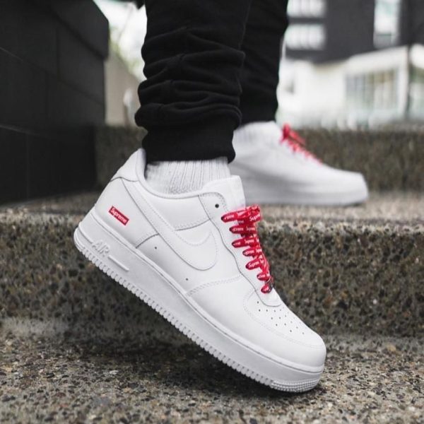 White Nike Air Force 1 sneakers with red laces stepping on a concrete outdoor stair, urban background, with black pants visible.