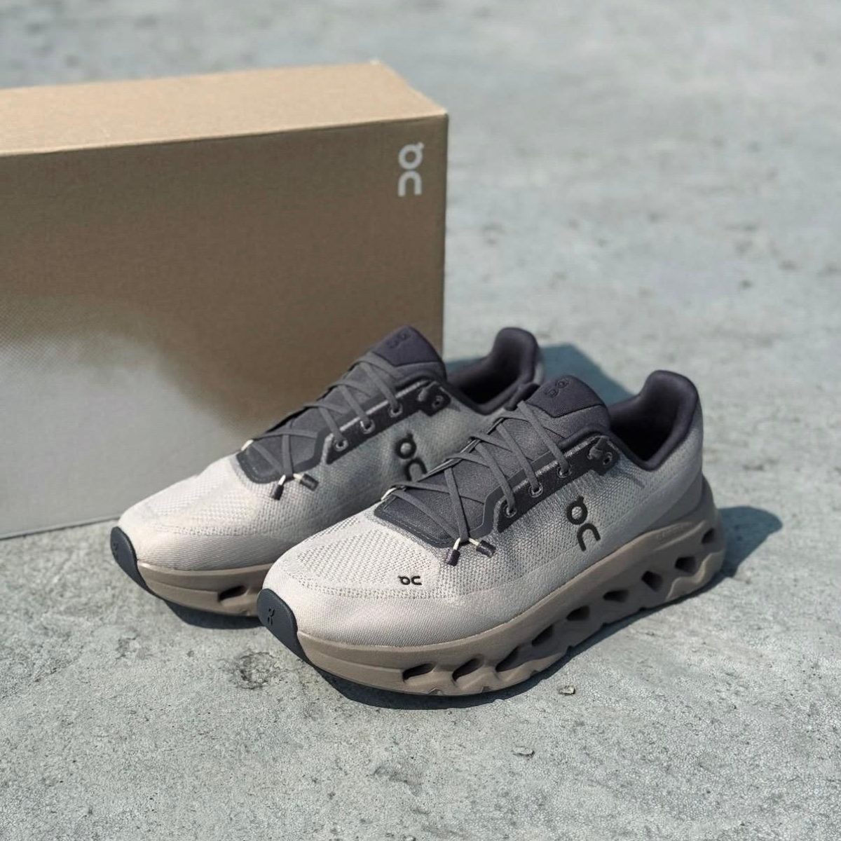 Gray On running shoes with cushioned, hollowed-out sole next to a brown box on a concrete surface, side view showing logo.