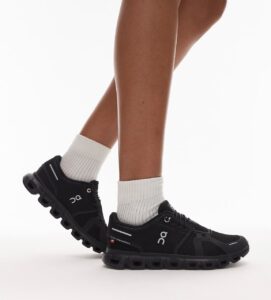Black athletic trail shoes with chunky, segmented soles worn with white crew socks on a person’s legs, logo visible on the sides.