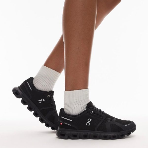 Black athletic trail shoes with chunky, segmented soles worn with white crew socks on a person’s legs, logo visible on the sides.