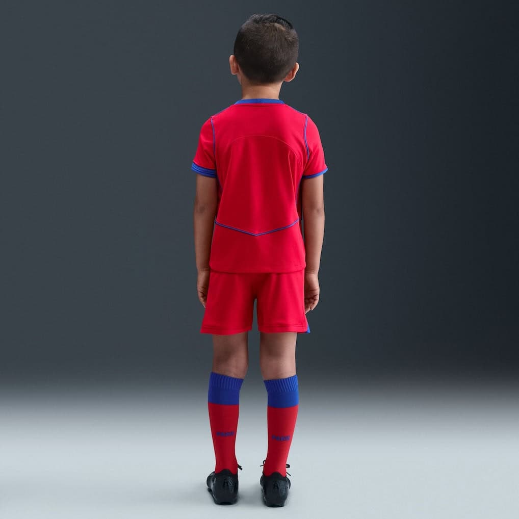 2q-1 | UAE SPORT STORE Back view of a young soccer player in a red kit with blue trim, standing against a dark gray studio backdrop.