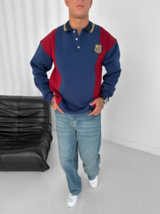 Man wearing a color-block polo with blue and maroon panels, FC Barcelona crest on the chest, jeans, and white sneakers in a studio setting.