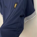 Navy polo shirt with a gold-embossed crest on the chest and striped white cuffs on the sleeves.