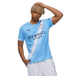 Young man wearing a Manchester City light-blue jersey, posing with hand on his head against a white background.