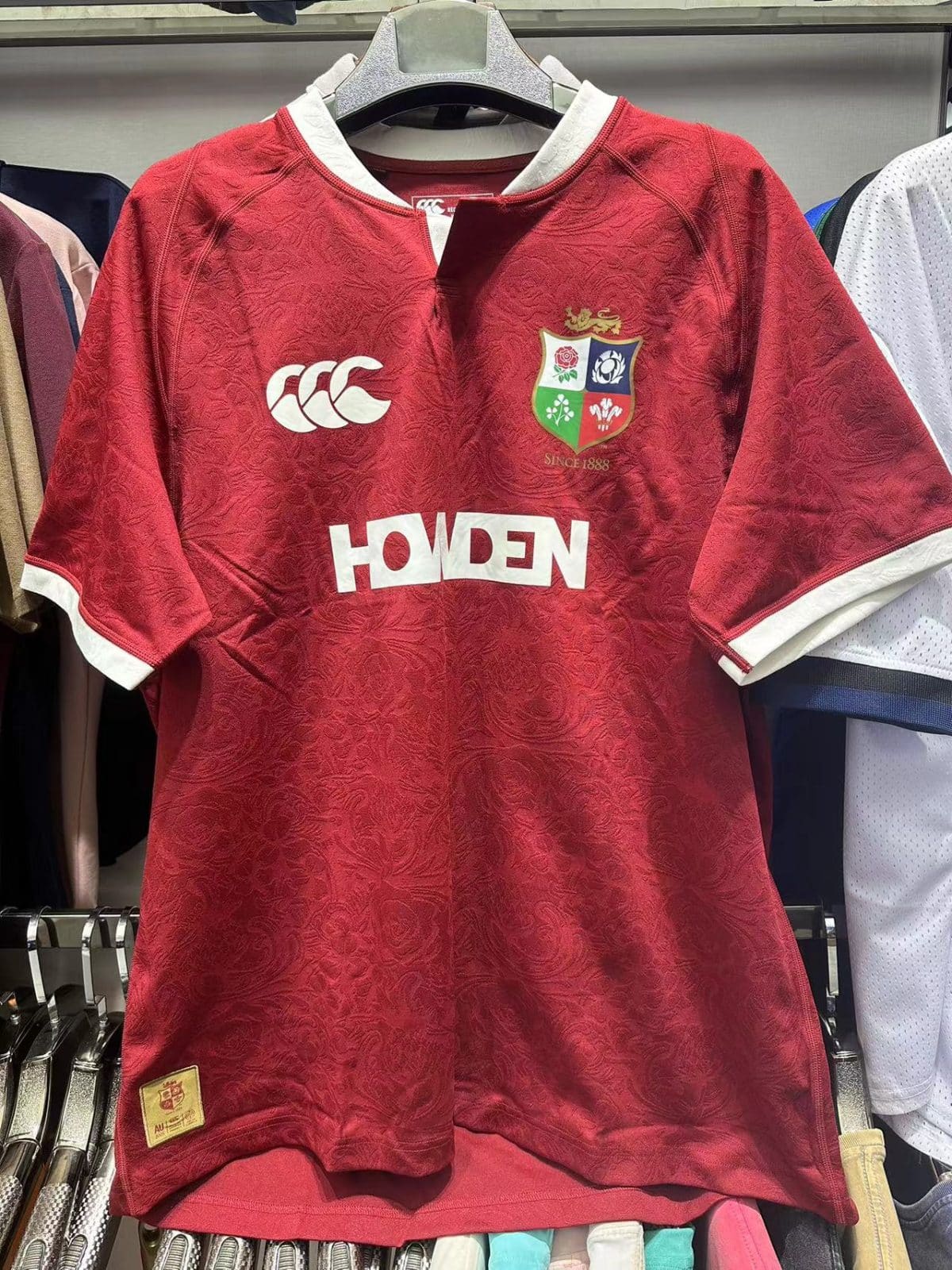 Red rugby jersey on a hanger with a crest emblem on the chest and white CCC logo; sponsor text 'HOWDEN' across the front.
