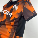 31b6a6c6_b0b3fc41-4d0e-484c-af9b-98f9a58376f6 | UAE SPORT STORE Close-up of an orange and black camouflage-patterned jersey with a white embroidered club crest and a gold star above it.