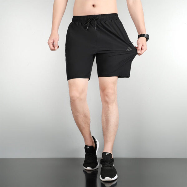 Man wearing black athletic shorts with a drawstring, standing with one hand pulling the fabric to show stretch, wearing black sneakers.