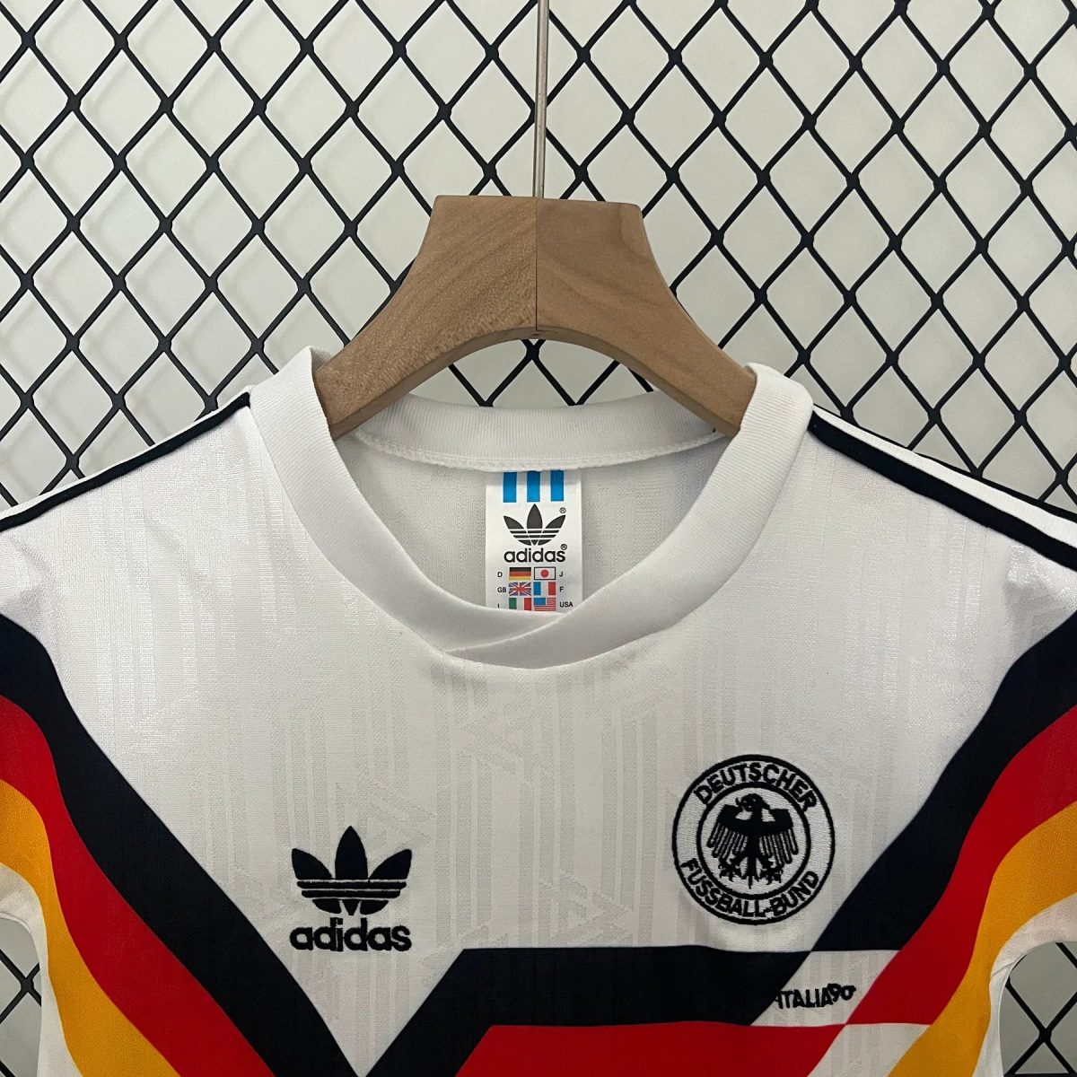 White Germany national football jersey on a wooden hanger, Adidas logo and DFB crest visible, with bold black, red, and yellow diagonal stripes in the foreground.