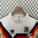 31d66ae9 | UAE SPORT STORE White Germany national football jersey on a wooden hanger, Adidas logo and DFB crest visible, with bold black, red, and yellow diagonal stripes in the foreground.