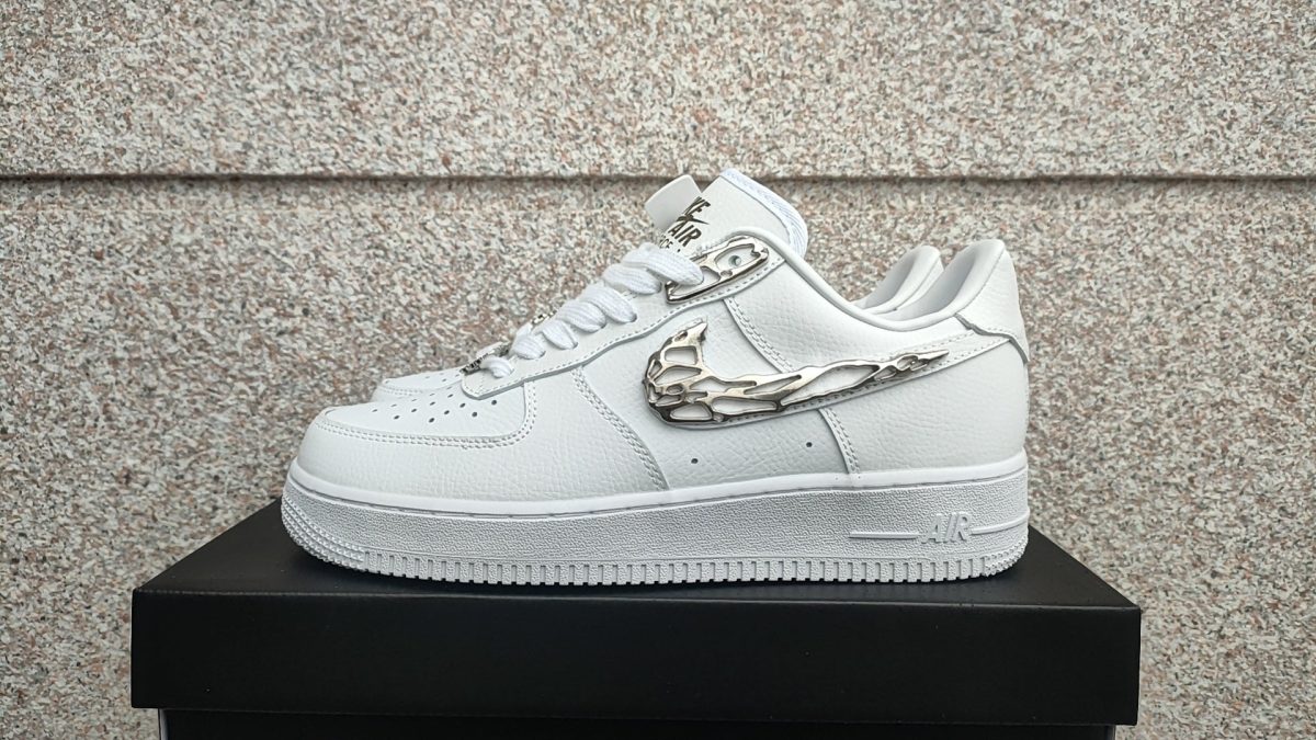 White leather sneaker with a metallic winged swoosh sits on a black box against a speckled stone wall.