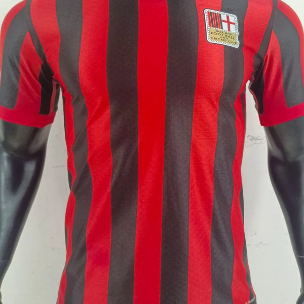 Red and black vertical striped soccer jersey on a black mannequin torso, with a small club crest badge on the left chest.