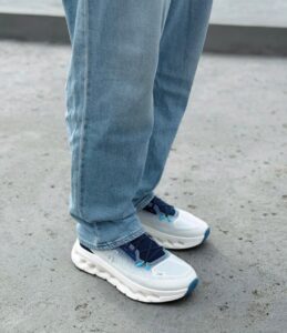 32e2adb7-459d-469a-b258-312151aa0bea | UAE SPORT STORE Person in light blue jeans standing on a gray pavement, wearing white and blue chunky sneakers with blue laces.