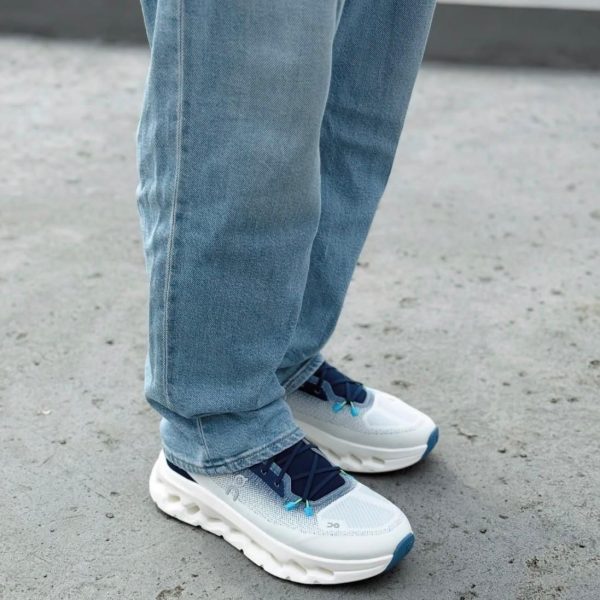 Person in light blue jeans standing on a gray pavement, wearing white and blue chunky sneakers with blue laces.
