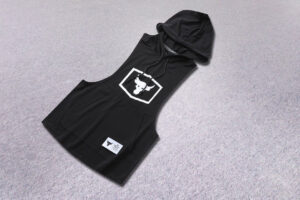 Black sleeveless hooded vest with a white square logo on the chest, laid on a gray carpeted floor, with a white label on the front pocket area.