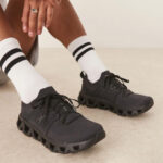 Person wearing black athletic sneakers with a chunky hollow sole and white socks with two black stripes on a light floor.