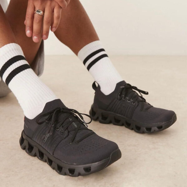 Person wearing black athletic sneakers with a chunky hollow sole and white socks with two black stripes on a light floor.