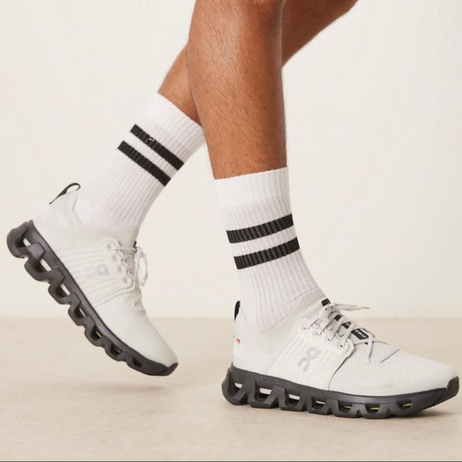 345e1768-c037-4c5b-904f-5f68a3086c2d White athletic sneakers with a distinctive black segmented sole, worn with white crew socks featuring two black stripes at the top edge.