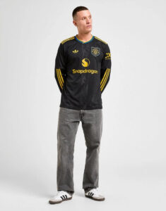 Male model wearing a black athletic jersey with yellow logos and stripes, gray jeans, and white sneakers against a light background.
