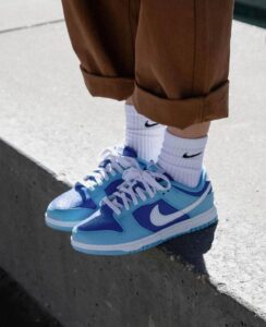 3500ee53-de16-4e66-878b-e23811eaff7a_e1b3233c-7bad-4b8b-8088-a413ea1c826c | UAE SPORT STORE Light blue Nike sneakers with white laces on white socks and rolled brown pants, standing on a concrete curb.