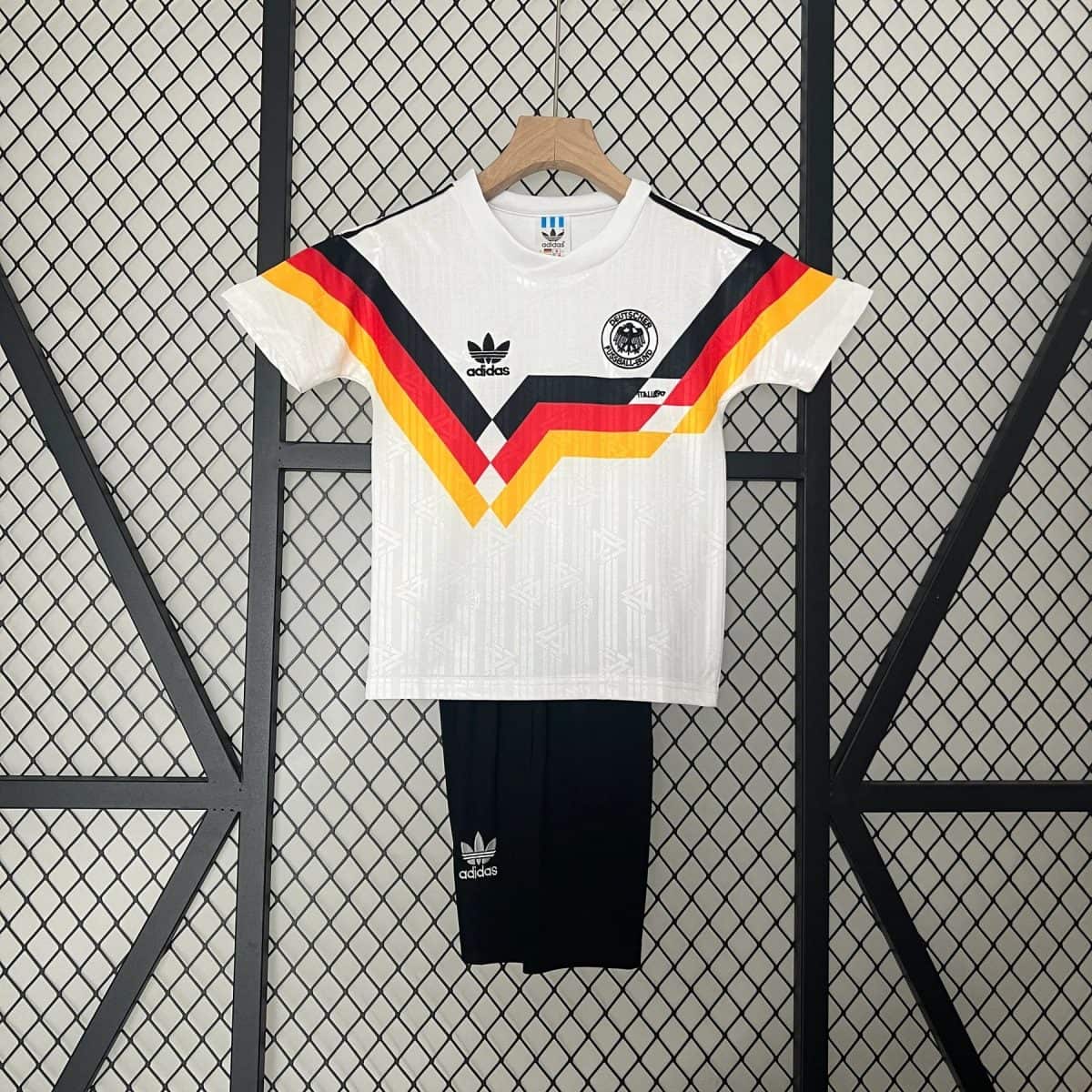 White Adidas Germany national team jersey on a wooden hanger, featuring black, red, and gold chevron stripes against a wire mesh backdrop.