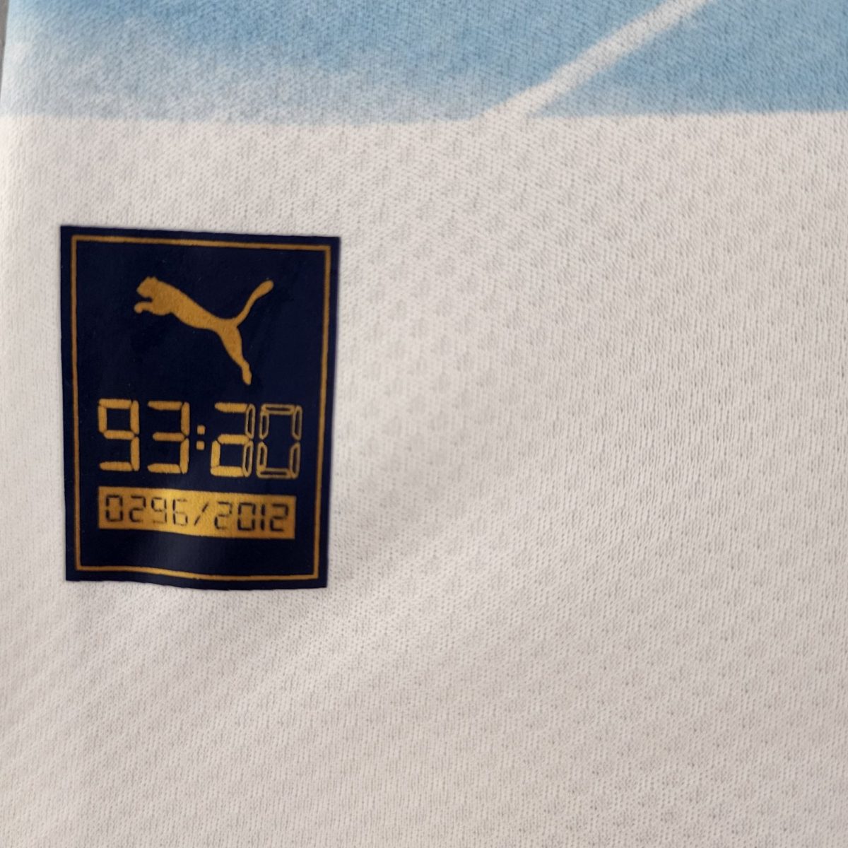 Close-up of a navy blue patch with a gold Puma logo stitched onto white fabric with a light blue stripe above it, likely a towel or garment tag.