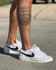 Leg tattoo shows clasped hands inside a laurel wreath with the text 'ROCKET FAMILIA', worn with white Nike sneakers with black swoosh.