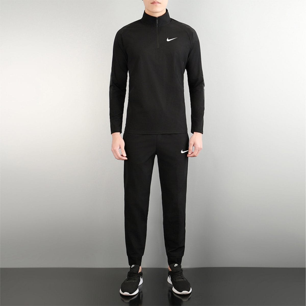 Person wearing a black Nike tracksuit with a white swoosh on the chest, standing against a gray background.