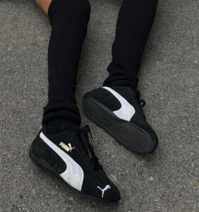 36c0d6bc-7454-4d67-a71f-4e235f993d86 | UAE SPORT STORE Person wearing black Puma sneakers with a white stripe, standing on asphalt with black socks/pants visible.