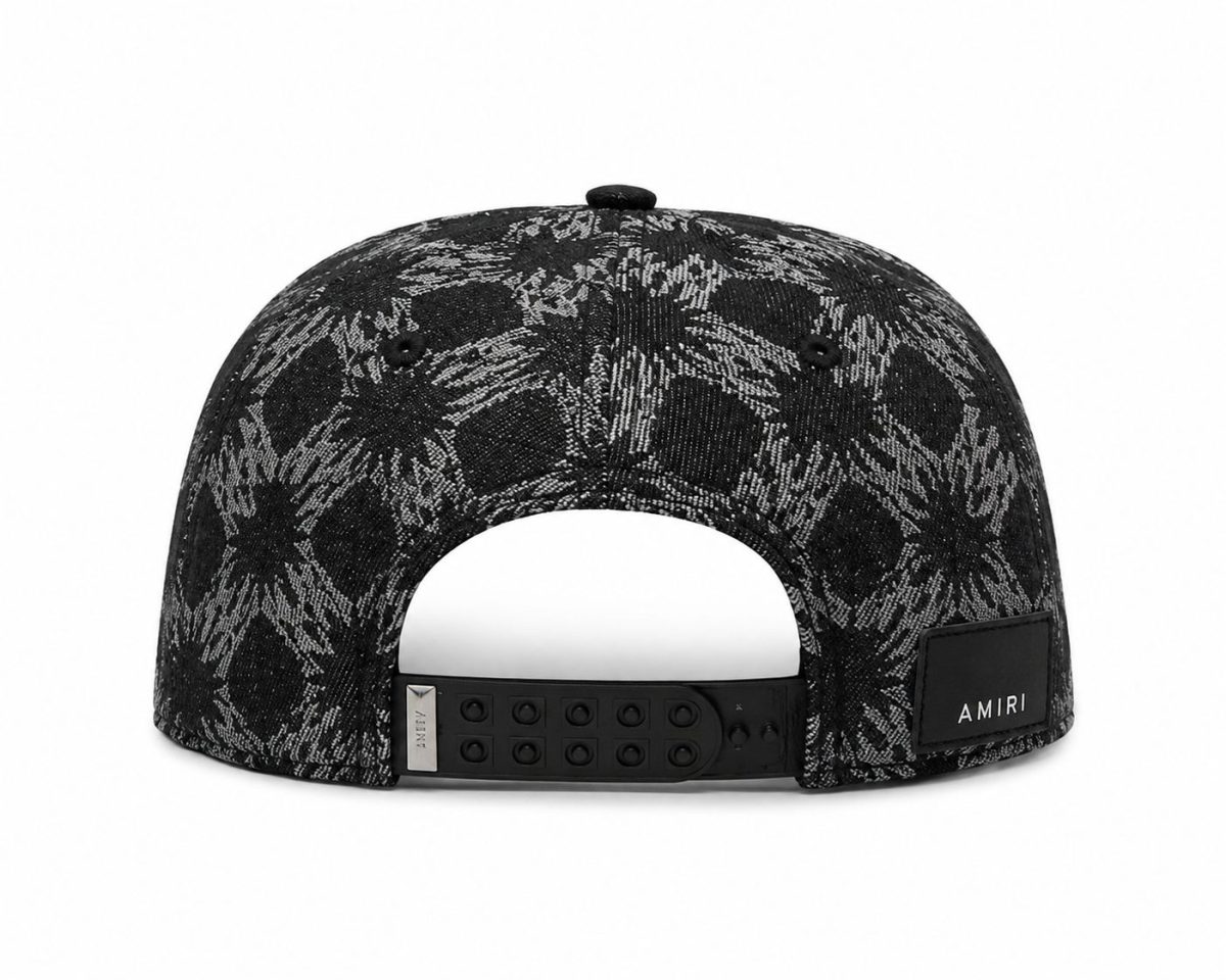 Back view of a black and gray patterned snapback cap with an adjustable plastic strap and an AMIRI label on the back.