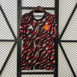 Manchester United football jersey with red-and-black flame pattern, Adidas logo on left chest and club crest on right, hanging on a hanger against a metal grid backdrop.
