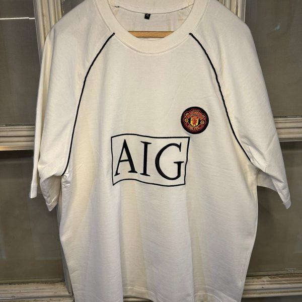 White Manchester United T-shirt on a hanger showing a large AIG sponsor logo and club crest on the chest area.