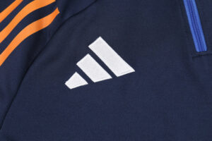 Close-up of a white Adidas three-bar logo on a navy athletic jacket with orange shoulder stripes and a blue zipper pull.
