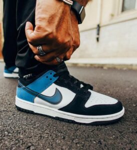 Close-up of a blue, white, and black Nike sneaker on a street, with a person adjusting the laces and wearing rings and a watch.