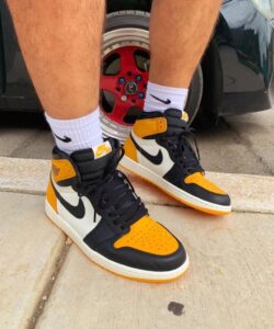 Pair of orange, black, and white Nike Air Jordan 1 sneakers with white socks on a concrete sidewalk next to a car wheel.