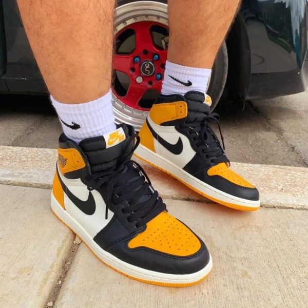 Pair of orange, black, and white Nike Air Jordan 1 sneakers with white socks on a concrete sidewalk next to a car wheel.