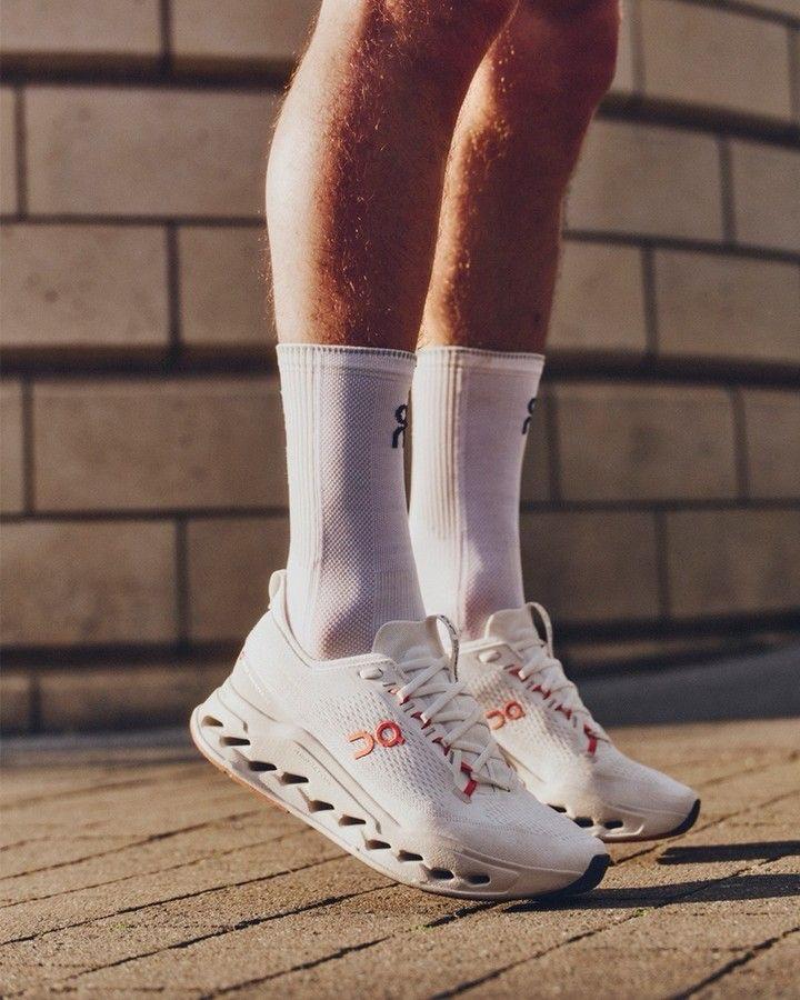 38cf51e9-e3c2-46d1-8864-57946b98af60 | UAE SPORT STORE Pair of white athletic shoes with a hollow-cut sole and white crew socks on a person standing on a brick sidewalk.