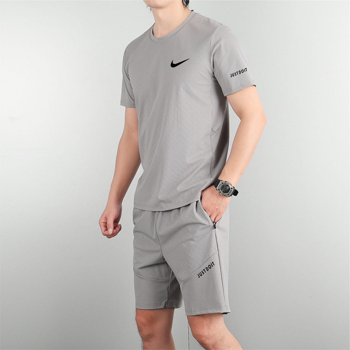 Man wearing a light gray athletic shirt with a black Nike swoosh on the chest and 'Just Do It' text visible on the sleeve/short leg edge, standing sideways with a watch on the wrist.