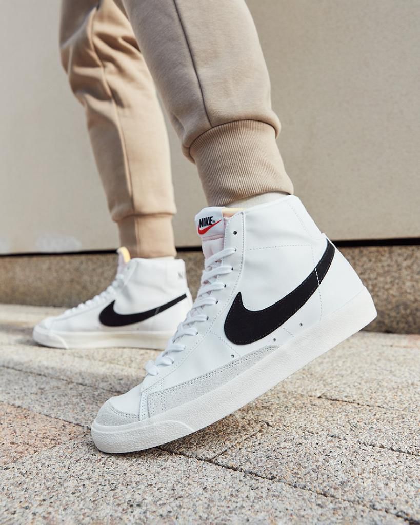 39c6134e-596d-4e81-aea1-2bae9ae2bee8 | UAE SPORT STORE White Nike high-top sneakers with a black swoosh, worn with beige joggers on a stone staircase.