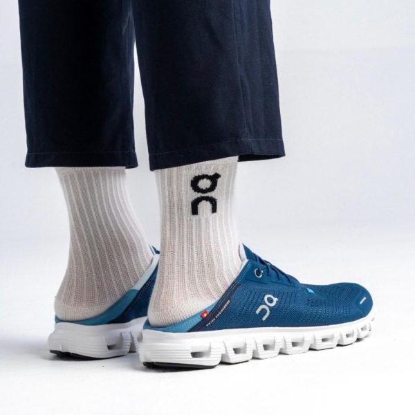 Side view of blue On running sneakers with a white chunky sole, paired with beige ribbed socks and dark cropped pants.