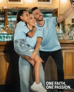 A family of three posing for a group selfie in a bar, wearing light blue soccer jerseys; the man holds the young girl while the woman smiles beside them.