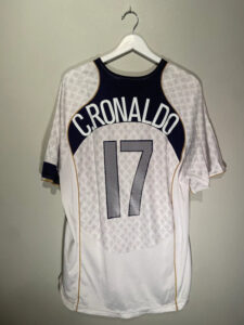 Back view of a white soccer jersey on a hanger, navy curved panel with gold trim reads 'CRONALDO' and the number 17