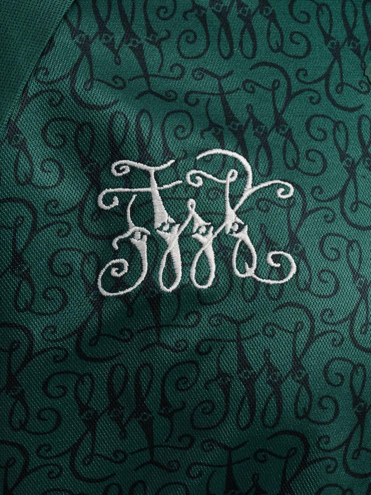 3Sj6xOouE9NsDt9 | UAE SPORT STORE Close-up of a white embroidered monogram on emerald green fabric with ornate swirls