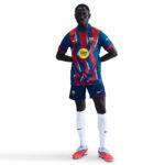 Soccer player standing in FC Barcelona kit with blue and red striped jersey, blue shorts, white socks, and pink cleats on a white background.