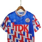 Blue geometric soccer jersey with red/white pattern, large TDK sponsor across the chest, Umbro logo and team crest, hung on a wooden hanger.