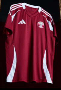 Maroon soccer jersey hanging on a rack with white Adidas stripes and a QFA crest on the chest area.0A sporty training jersey with maroon color and white side panels.