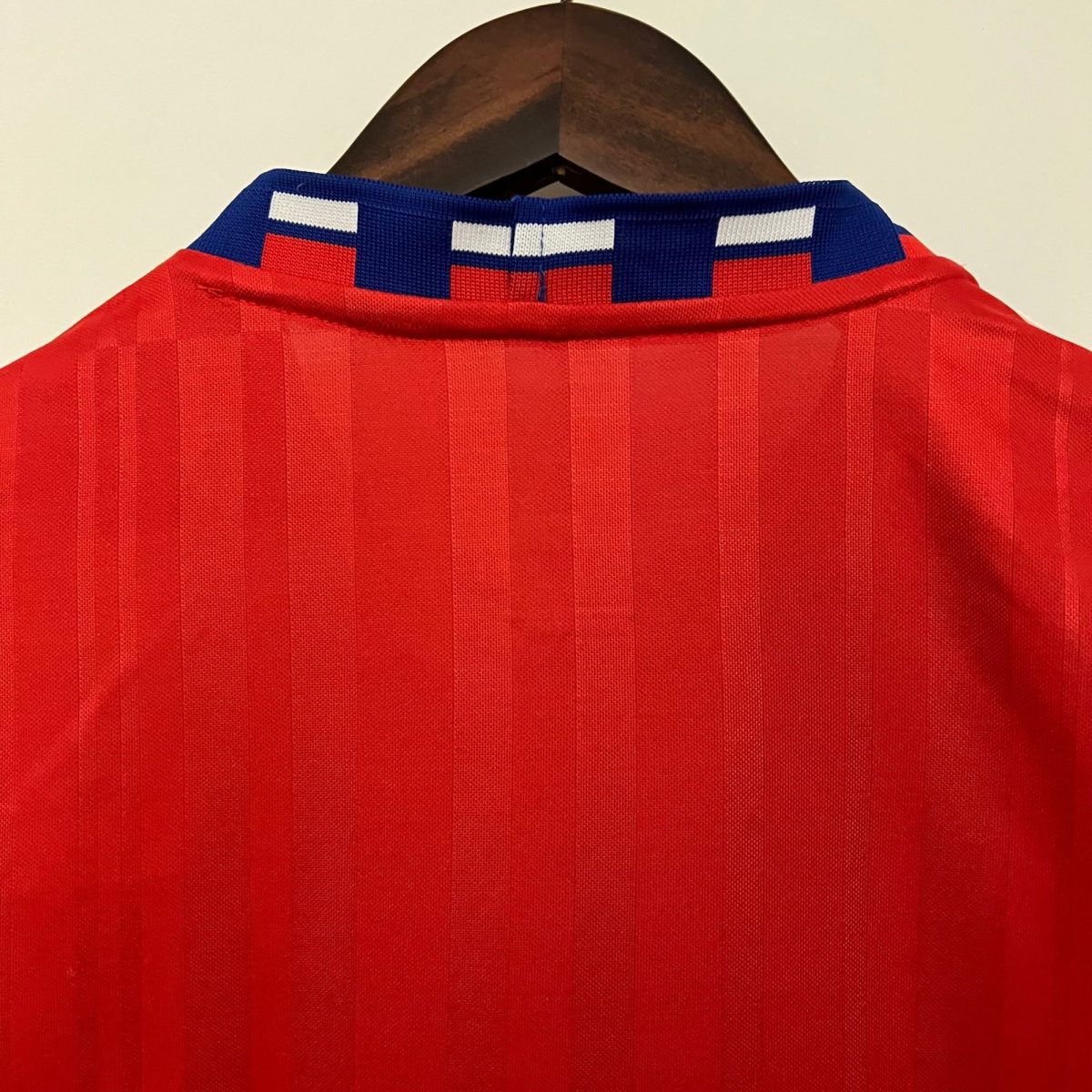 Back view of a red sports jersey on a wooden hanger, showing a blue collar with white blocks at the neck.